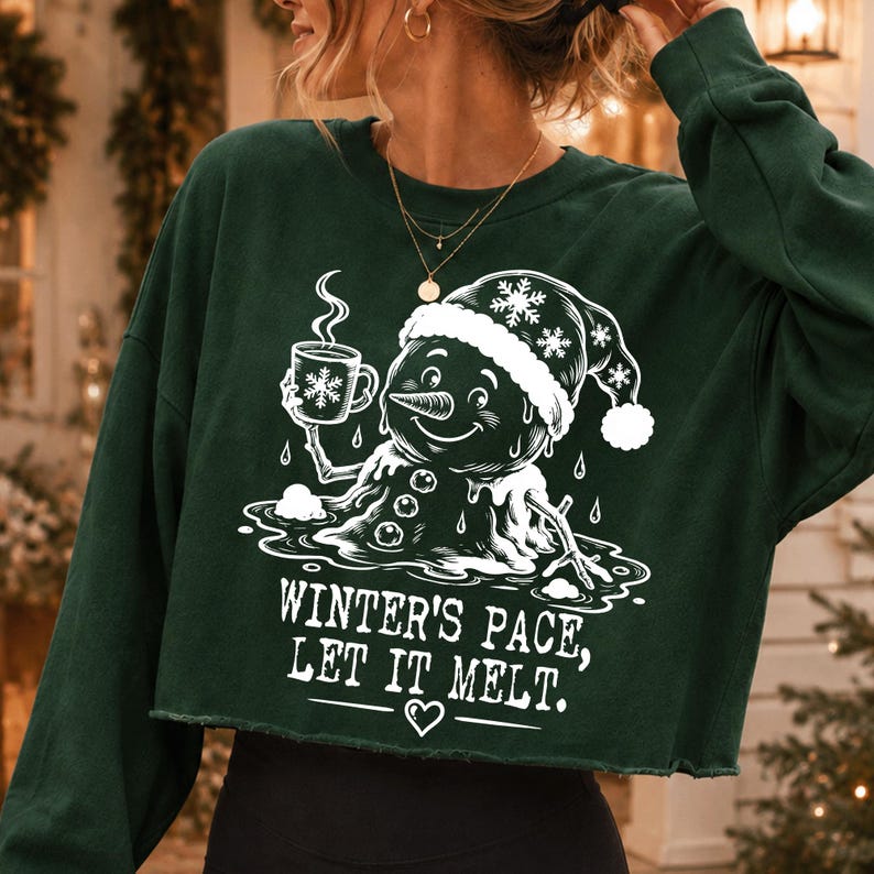 May include: A dark green long-sleeve cropped sweatshirt featuring a white graphic of a melting snowman wearing a Santa hat and holding a mug with a snowflake design. The text reads: WINTER'S PACE, LET IT MELT. with a small heart icon below the text.
