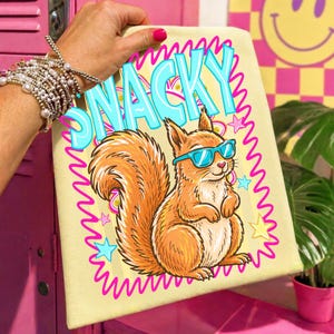 May include: A light yellow canvas with a cartoon squirrel wearing blue sunglasses. The word "SNACKY" is in large, light blue letters outlined in pink. The squirrel is orange with a fluffy tail, surrounded by pink stars and a pink wavy border.