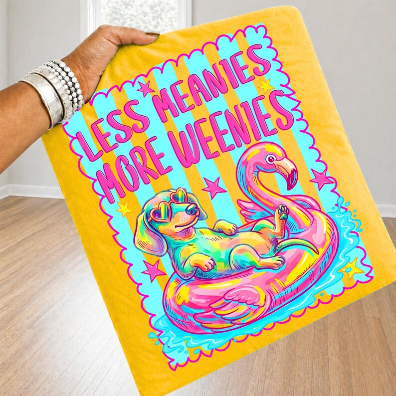 May include: A yellow throw blanket featuring a colorful graphic of a dachshund wearing heart-shaped sunglasses while floating on a pink flamingo pool float. The text reads LESS MEANIES MORE WEENIES in bright pink letters surrounded by small stars.