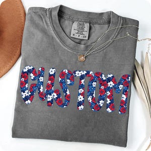 K&ouml;nnte beinhalten: Ein graues Comfort Colors T-Shirt mit dem Wort "CUSTOM" in einem floralen Design aus roten, wei&szlig;en und blauen Blumen. Eine silberne Halskette mit Anh&auml;nger liegt auf dem Shirt. Ein brauner Hut ist oben links sichtbar.