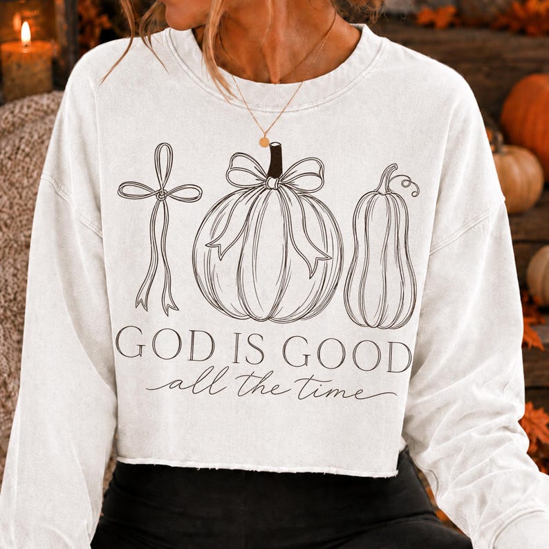 K&ouml;nnte beinhalten: Ein wei&szlig;es Langarm-Sweatshirt mit einer schwarzen Strichzeichnung eines Schleifenbandes, eines K&uuml;rbisses mit Schleife und eines Zierk&uuml;rbisses. Unter den Abbildungen steht der Text "GOD IS GOOD all the time" in einer Kombination aus Serif- und Schreibschrift.