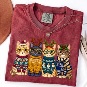 May include: A maroon t-shirt featuring a graphic of four cats wearing holiday sweaters and glasses. The first cat wears a Santa hat, the second has reindeer antlers, the third has mistletoe, and the fourth has holly. The Comfort Colors tag is visible.