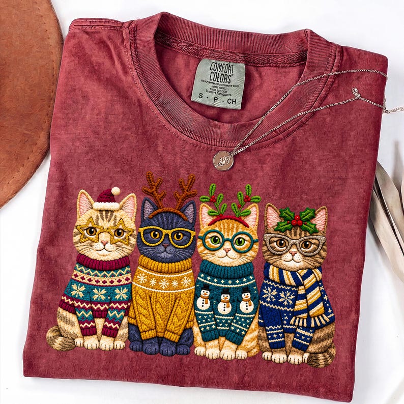 May include: A maroon t-shirt featuring a graphic of four cats wearing holiday sweaters and glasses. The first cat wears a Santa hat, the second has reindeer antlers, the third has mistletoe, and the fourth has holly. The Comfort Colors tag is visible.