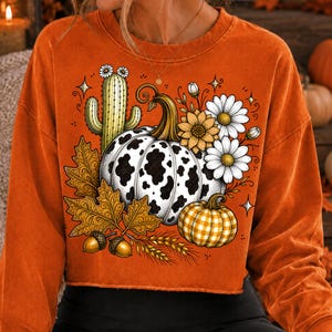 Puede incluir: Una sudadera naranja de manga larga con un estampado gr&aacute;fico de una calabaza con manchas de vaca, una calabaza de cuadros amarillos, un cactus verde con flores blancas, hojas de oto&ntilde;o marrones, bellotas, espigas de trigo y margaritas blancas.