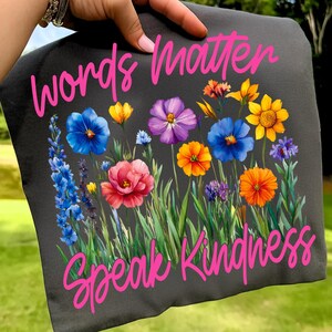 Puede incluir: Camiseta gris oscuro con un dise&ntilde;o floral vibrante. El dise&ntilde;o incluye flores azules, moradas, naranjas y amarillas. Las palabras "Words Matter" y "Speak Kindness" est&aacute;n escritas en rosa en la parte superior e inferior.