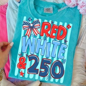 Puede incluir: Camiseta turquesa Comfort Colors con un dise&ntilde;o patri&oacute;tico. El dise&ntilde;o presenta las palabras "RED WHITE & 250" en rojo y azul, junto con un lazo, estrellas y fuegos artificiales. La camiseta es de cuello redondo.