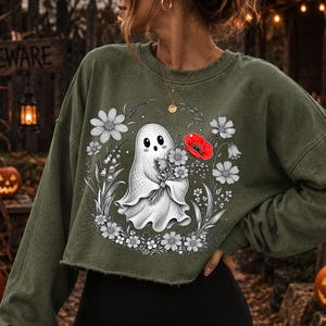 K&ouml;nnte beinhalten: Ein olivgr&uuml;nes Langarm-Sweatshirt mit einem Grafikdruck eines wei&szlig;en Geistes, der einen Blumenstrau&szlig; h&auml;lt. Das Design zeigt eine kreisf&ouml;rmige Anordnung von schwarz-wei&szlig;en Strichzeichnungen von Blumen mit einer einzelnen leuchtend roten Mohnblume.