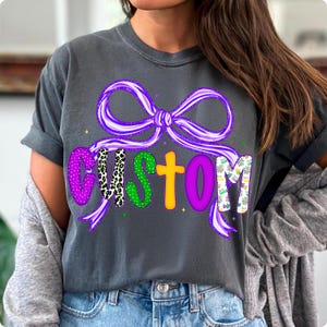 May include: Dark gray t-shirt with a purple bow and the word "CUSTOM" in colorful letters. The letters have various patterns, including polka dots and animal print. The shirt is a casual style.