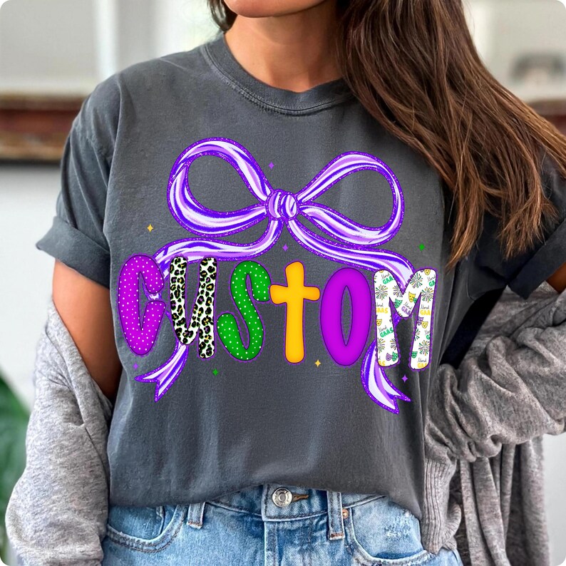 May include: Dark gray t-shirt with a purple bow and the word "CUSTOM" in colorful letters. The letters have various patterns, including polka dots and animal print. The shirt is a casual style.