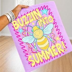K&ouml;nnte beinhalten: Eine hellviolette Decke mit den Worten "BUZZIN INTO SUMMER!" in Gelb. Die Decke zeigt eine Cartoon-Biene mit gelben und schwarzen Streifen, blauen Fl&uuml;geln und blauen und gelben Akzenten. Rosa Sterne und ein rosa gewellter Rand umgeben den Text und die Biene.