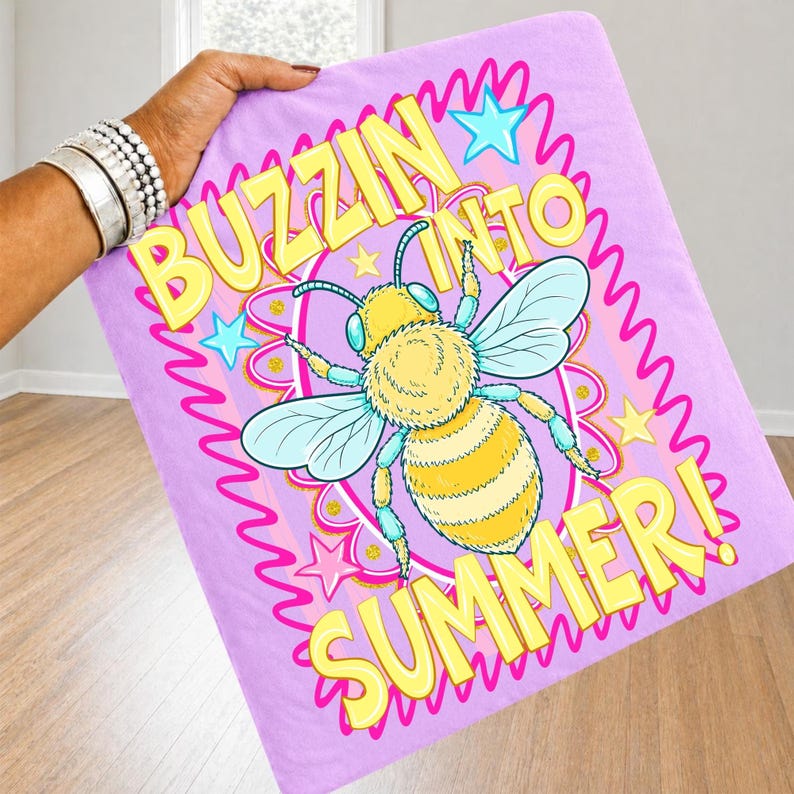 K&ouml;nnte beinhalten: Eine hellviolette Decke mit den Worten "BUZZIN INTO SUMMER!" in Gelb. Die Decke zeigt eine Cartoon-Biene mit gelben und schwarzen Streifen, blauen Fl&uuml;geln und blauen und gelben Akzenten. Rosa Sterne und ein rosa gewellter Rand umgeben den Text und die Biene.