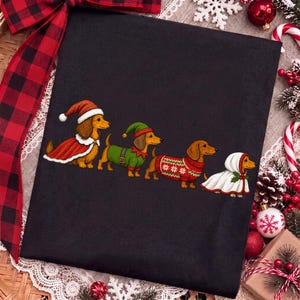 May include: A black t-shirt featuring a graphic of four brown dachshunds in a row. The first wears a Santa hat and cape, the second an elf outfit, the third a red patterned holiday jumper, and the fourth a white hooded cloak with holly berries.