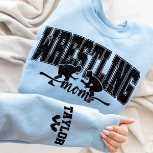 May include: Light blue sweatshirt with the word "WRESTLING" in large, distressed black letters. Below the word are two black silhouettes of wrestlers and the word "mom". The sleeve has the name "TAYLOR" in black.
