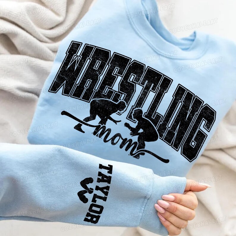 May include: Light blue sweatshirt with the word "WRESTLING" in large, distressed black letters. Below the word are two black silhouettes of wrestlers and the word "mom". The sleeve has the name "TAYLOR" in black.