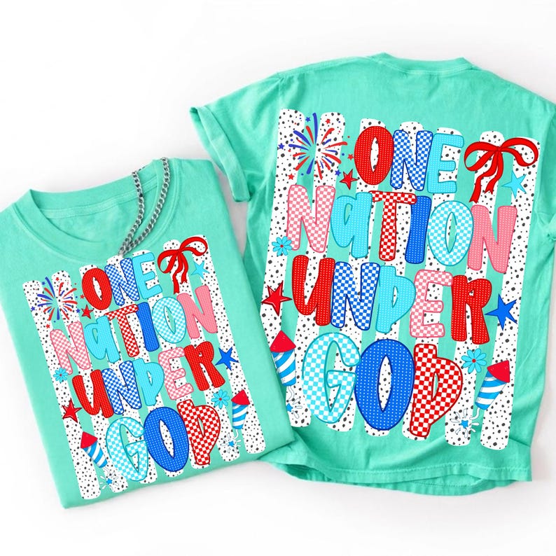 Puede incluir: Dos camisetas verde menta con el texto "ONE NATION UNDER GOD" en letras de burbujas rojas, azules y rosas. El dise&ntilde;o incluye fuegos artificiales, estrellas y un lazo. Las camisetas son probablemente para un d&iacute;a festivo patri&oacute;tico.