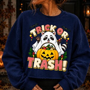 May include: A navy blue long-sleeve sweatshirt featuring a graphic of a raccoon dressed as a ghost, holding a jack-o'-lantern bucket filled with sweets. The text reads TRICK OR TRASH! with red and white chequered accents and small black bats.