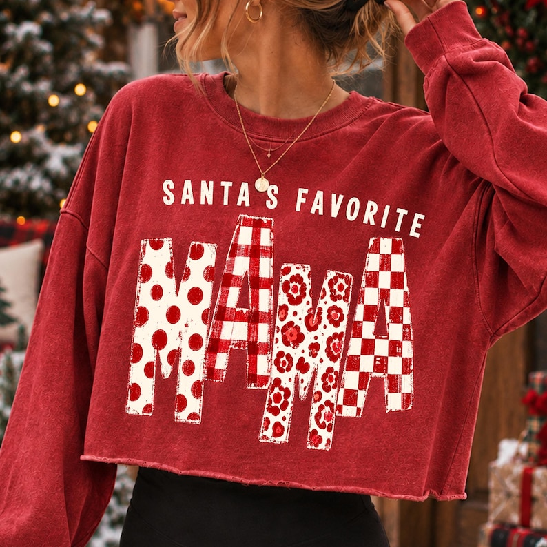 May include: A red long-sleeve cropped sweatshirt featuring the white text SANTA'S FAVORITE above the word MAMA. The letters in MAMA are filled with different red and white patterns, including polka dots, plaid, floral prints, and a checkerboard design.