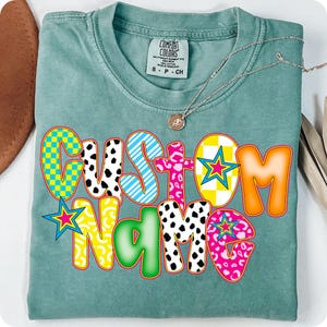 May include: A teal Comfort Colors t-shirt with the words "CUSTOM NAME" in a colorful, playful font. The letters feature various patterns, including checkered, striped, and animal print designs. A silver necklace with a pendant rests on the shirt.