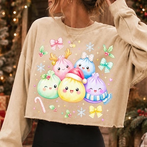 May include: A beige long-sleeve cropped sweatshirt featuring a graphic print of five colourful, smiling dumpling characters. The dumplings are decorated with holiday elements including a Santa hat, reindeer antlers, a scarf, and bows, surrounded by snowflakes.