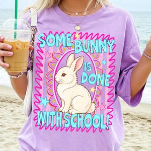 Może przedstawiać: Liliowa koszulka z napisem "SOME BUNNY IS DONE WITH SCHOOL!" w kolorze turkusowym. Biały kr&oacute;lik jest umieszczony wewnątrz r&oacute;żowo-ż&oacute;łtego owalu. Koszulka ma kr&oacute;tkie rękawy.