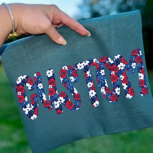K&ouml;nnte beinhalten: T&uuml;rkisfarbener Stoff mit dem Wort "CUSTOM" in gro&szlig;en, fetten Buchstaben. Die Buchstaben sind mit einem Blumenmuster in Rot, Wei&szlig; und Blau gef&uuml;llt. Der Stoff wird von einer Hand gehalten, die das individuelle Design zeigt.