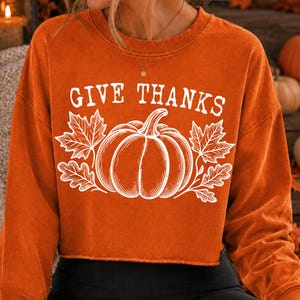 Puede incluir: Una sudadera de manga larga y cuello redondo en color naranja quemado con dobladillo sin rematar. El frente presenta un estampado gr&aacute;fico blanco de una calabaza flanqueada por hojas de arce y roble, con el texto "GIVE THANKS" en blanco.