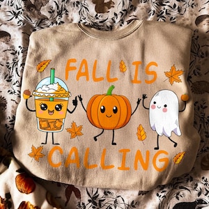 May include: A beige crewneck sweatshirt featuring a graphic print with the orange text FALL IS CALLING. The design includes a cartoon iced coffee, a smiling orange pumpkin, and a white ghost, all surrounded by small orange and yellow autumn leaves.