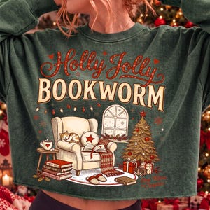 Op de afbeelding: Een donkergroene sweater met ronde hals en een grafische print met de tekst Holly Jolly BOOKWORM. Het ontwerp toont een comfortabele fauteuil met een slapende kat, een open boek, een kerstboom, ingepakte cadeaus en een raam met sneeuw.