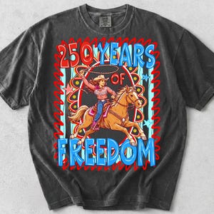 May include: Dark gray t-shirt featuring a graphic of a cowgirl riding a horse, with the text "250 YEARS OF FREEDOM" in blue and red. The design includes stars and a lasso, celebrating a theme of freedom.