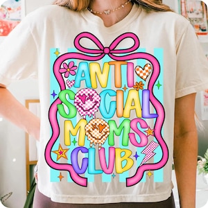 K&ouml;nnte beinhalten: Cremefarbenes T-Shirt mit dem Text "ANTI SOCIAL MOMS CLUB" in farbenfrohen, verspielten Buchstaben. Das Design umfasst ein rosa Band, Blumen, Herzen, Sterne und Blitze. Das Shirt hat einen Rundhalsausschnitt und kurze &Auml;rmel.