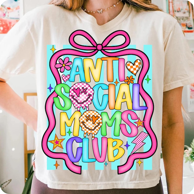 K&ouml;nnte beinhalten: Cremefarbenes T-Shirt mit dem Text "ANTI SOCIAL MOMS CLUB" in farbenfrohen, verspielten Buchstaben. Das Design umfasst ein rosa Band, Blumen, Herzen, Sterne und Blitze. Das Shirt hat einen Rundhalsausschnitt und kurze &Auml;rmel.