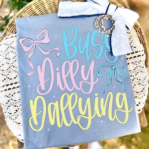 Puede incluir: Una camiseta azul claro con las palabras "Busy Dilly Dallying" en escritura azul, rosa y amarilla. La camiseta presenta gr&aacute;ficos de lazos y est&aacute; adornada con un lazo blanco y pulseras.
