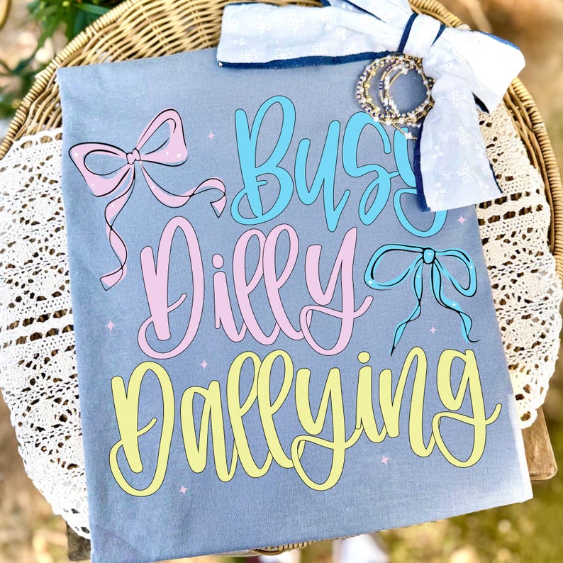 Puede incluir: Una camiseta azul claro con las palabras "Busy Dilly Dallying" en escritura azul, rosa y amarilla. La camiseta presenta gr&aacute;ficos de lazos y est&aacute; adornada con un lazo blanco y pulseras.