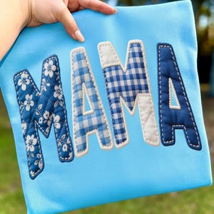 May include: A light blue fabric item with the word "MAMA" appliqued in various blue patterns. The "M" features a floral design, the "A"s have gingham, and the final "A" is solid blue. White stitching outlines each letter.
