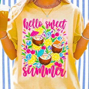 May include: A pale yellow t-shirt featuring a vibrant graphic print with the text "hello sweet summer" in bright pink script. The design includes illustrations of halved coconuts with colorful cocktail umbrellas, lemon slices, cherries, and blue ice cubes.