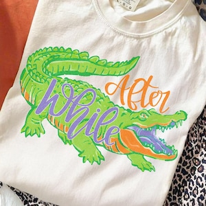 Pu&ograve; includere: T-shirt color crema con una grande illustrazione di un alligatore verde con accenti arancioni e viola. Le parole "While" e "After" sono scritte in corsivo viola e arancione sull'alligatore. La maglietta ha uno scollo tondo.