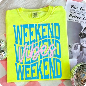 K&ouml;nnte beinhalten: Ein neongelbes T-Shirt mit den Worten "WEEKEND VIBES WEEKEND" in blauen und rosa Buchstaben. Das Shirt ist von der Marke Comfort Colors und die Gr&ouml;&szlig;e ist L-G. Das Shirt wird auf einer rosa-wei&szlig; karierten Oberfl&auml;che pr&auml;sentiert.