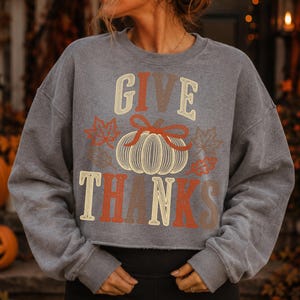 Puede incluir: Una sudadera gris carb&oacute;n de manga larga con el texto GIVE THANKS en una fuente retro. El dise&ntilde;o incluye una calabaza central dibujada con l&iacute;neas y un lazo rojo, rodeada de varias ilustraciones de hojas de oto&ntilde;o en tonos naranja y marr&oacute;n.