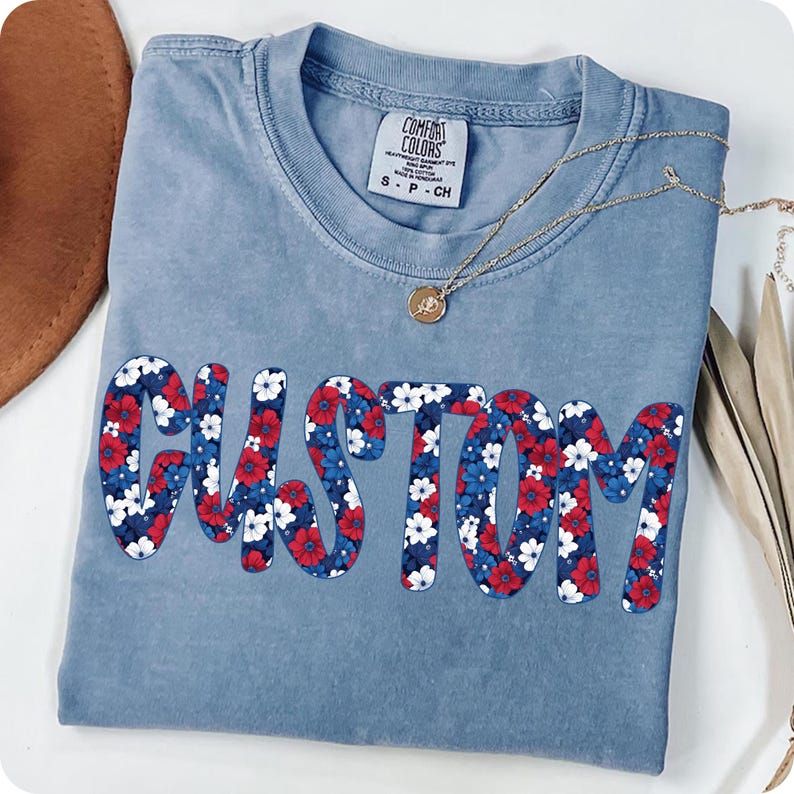 K&ouml;nnte beinhalten: Hellblaues Comfort Colors T-Shirt mit dem Wort "CUSTOM" in einem Blumenmuster aus Rot, Wei&szlig; und Blau. Eine goldene Halskette mit Anh&auml;nger liegt &uuml;ber dem Shirt.