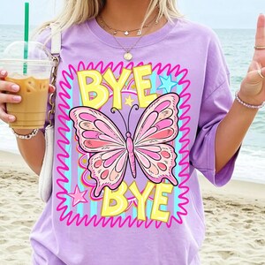 May include: A lilac t-shirt with a large butterfly graphic in pink and peach. The words "BYE BYE" are in yellow with a pink outline, set against a blue and pink background. The shirt has short sleeves and a relaxed fit.
