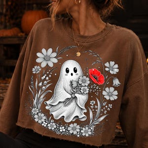 K&ouml;nnte beinhalten: Ein braunes bauchfreies Sweatshirt mit einem Grafikdruck eines wei&szlig;en Geistes, der einen Blumenstrau&szlig; h&auml;lt. Das Design zeigt schwarz-wei&szlig;e Strichzeichnungen von Blumen um den Geist herum, mit einer einzelnen leuchtend roten Mohnblume als Blickfang.