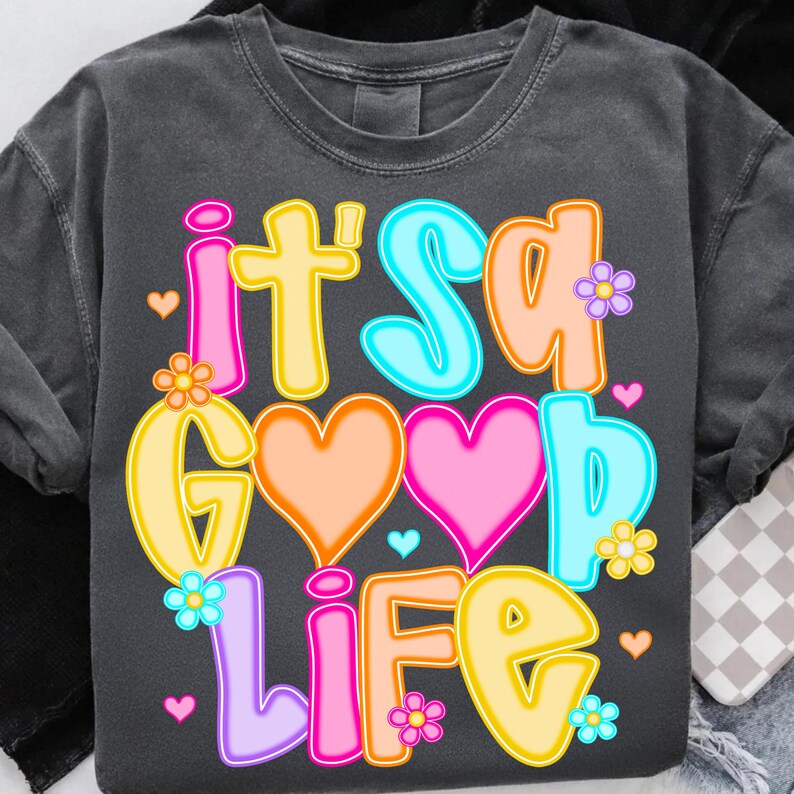 Pode incluir: T-shirt cinza escuro com a frase "It's a Good Life" em letras coloridas. O design inclui cora&ccedil;&otilde;es e flores em tons de rosa, laranja, amarelo e azul. Uma camiseta gr&aacute;fica casual e alegre.