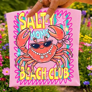 May include: Pink t-shirt featuring a crab graphic wearing sunglasses and a pearl necklace. The text "SALT MOM'S BEACH CLUB" is in yellow and blue. The crab is coral-coloured with pink accents. The background is a garden.