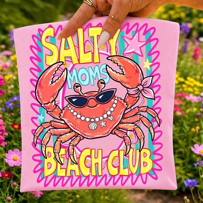 May include: Pink t-shirt featuring a crab graphic wearing sunglasses and a pearl necklace. The text "SALT MOM'S BEACH CLUB" is in yellow and blue. The crab is coral-coloured with pink accents. The background is a garden.