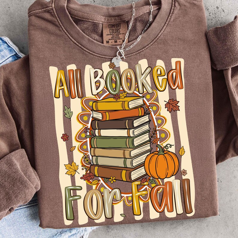 Op de afbeelding: Een bruin sweatshirt met lange mouwen en een grafische print van een stapel boeken en een pompoen. De tekst luidt All Booked For Fall. Het ontwerp bevat herfstbladeren en bloemen in tinten oranje, geel, groen en bruin op een gestreepte achtergrond.