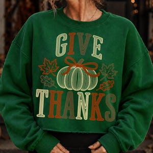 Puede incluir: Una sudadera de manga larga de color verde oscuro con el texto GIVE THANKS en una fuente de estilo retro de varias capas. Centrado entre las palabras hay un gr&aacute;fico de una calabaza con un lazo, rodeado de hojas de arce y roble.
