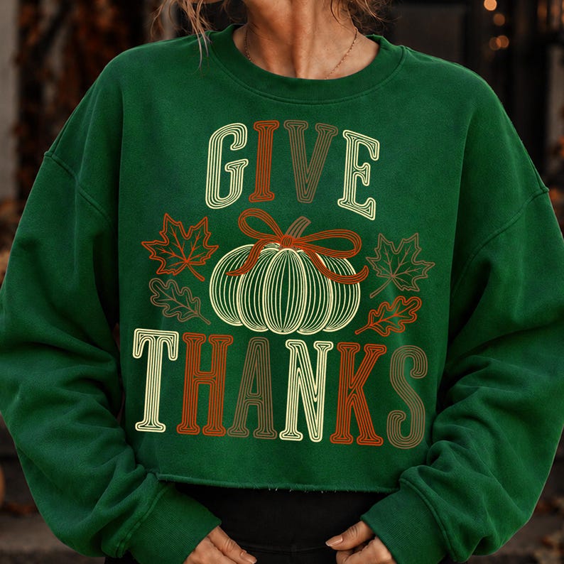 Puede incluir: Una sudadera de manga larga de color verde oscuro con el texto GIVE THANKS en una fuente de estilo retro de varias capas. Centrado entre las palabras hay un gr&aacute;fico de una calabaza con un lazo, rodeado de hojas de arce y roble.