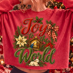 Może przedstawiać: Czerwona bluza z okrągłym dekoltem z nadrukiem graficznym z napisem Joy to the World. Wz&oacute;r przedstawia scenę narodzin, białe jagnię, gwiazdy betlejemskie, liście ostrokrzewu, czerwone jagody oraz czerwono-białą kokardę w kratkę.