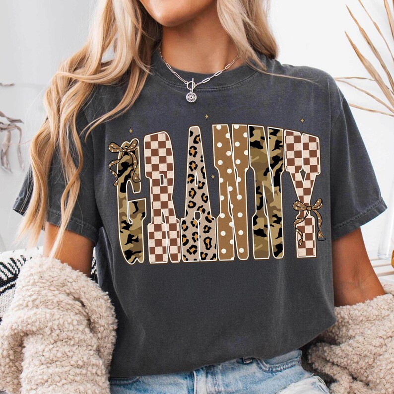 May include: A charcoal grey short-sleeve t-shirt featuring the word GRANNY printed in large, stylised letters. The letters incorporate various patterns including brown and white checkerboard, leopard print, camouflage, and white polka dots.