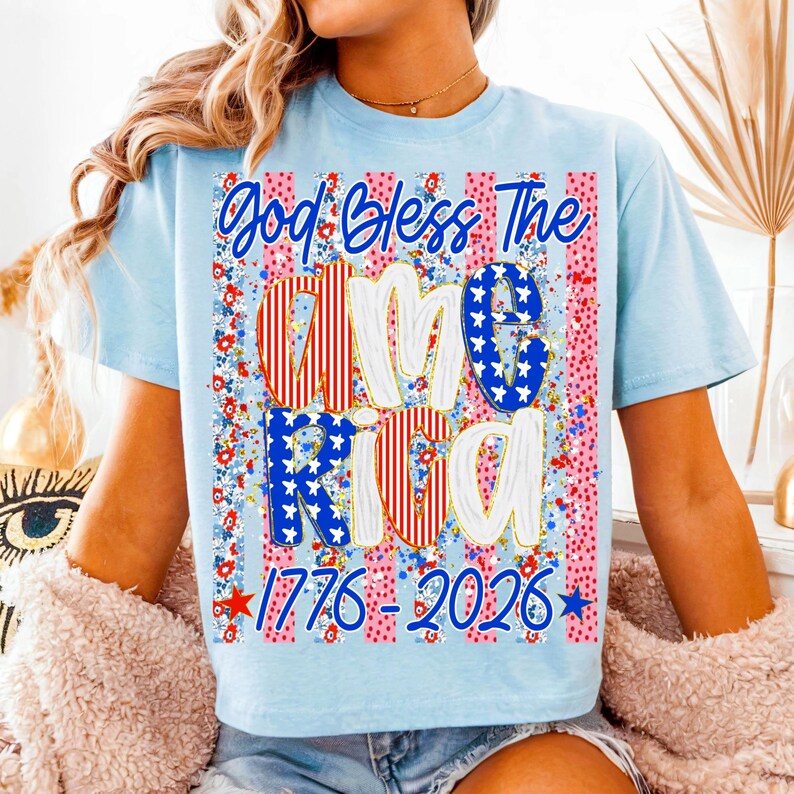 Puede incluir: Camiseta azul claro con el texto "God Bless The America" en un dise&ntilde;o patri&oacute;tico. La palabra "America" est&aacute; rellena de rayas rojas y blancas y estrellas azules. La camiseta tambi&eacute;n tiene las fechas "1776-2026".